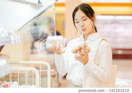 A young woman compares white eggs and brown eggs in the supermarket's egg section to choose one. A young woman compares white eggs and brown eggs in the supermarket's egg section to choose one. 142203519