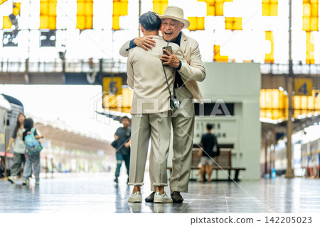 Two senior LGBTQ men in cream suits embrace warmly at train station with yellow lanterns overhead Two senior LGBTQ men in cream suits embrace warmly at train station with yellow lanterns overhead 142205023