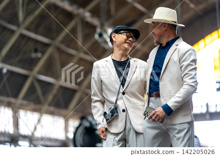 Two senior LGBTQ men in cream suits share warm smiles and laughter under station's wooden beams Two senior LGBTQ men in cream suits share warm smiles and laughter under station's wooden beams 142205026