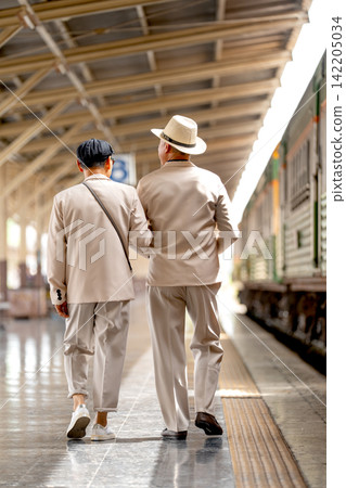 Two senior LGBTQ men in cream suits walk side by side along train station platform, one with straw hat Two senior LGBTQ men in cream suits walk side by side along train station platform, one with straw hat 142205034