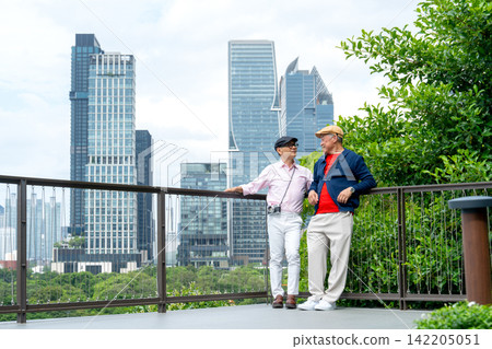 Two senior LGBTQ men stand on modern balcony, one holds camera as they gaze at city skyline through lush greenery Two senior LGBTQ men stand on modern balcony, one holds camera as they gaze at city skyline through lush greenery 142205051