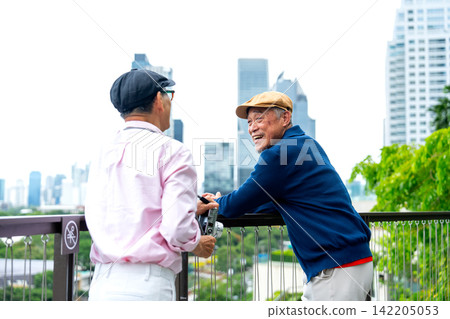 Two senior LGBTQ men share joyful moment on modern balcony with city skyline and lush greenery backdrop 142205053