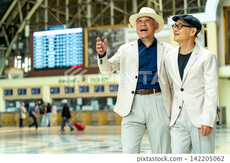 Two joyful senior men in matching white suits laugh together at train station, one pointing excitedly toward departure board. Two joyful senior men in matching white suits laugh together at train station, one pointing excitedly toward departure board. 142205062