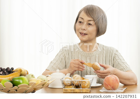 A senior woman eating breakfast bread in the living room. 142207931