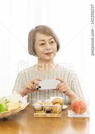 一位老婦人在客廳一邊吃早餐麵包一邊使用智慧型手機。 一位老婦人在客廳一邊吃早餐麵包一邊使用智慧型手機。 142208257