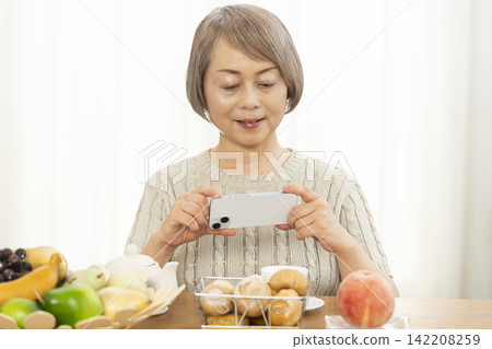 A senior woman eating breakfast bread while using her smartphone in the living room. A senior woman eating breakfast bread while using her smartphone in the living room. 142208259