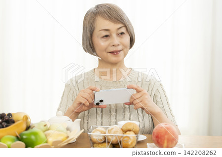 一位老婦人在客廳一邊吃早餐麵包一邊使用智慧型手機。 一位老婦人在客廳一邊吃早餐麵包一邊使用智慧型手機。 142208262