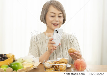 A senior woman eating breakfast bread while using her smartphone in the living room. A senior woman eating breakfast bread while using her smartphone in the living room. 142208264