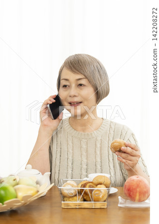 A senior woman eating breakfast bread while using her smartphone in the living room. 142208272