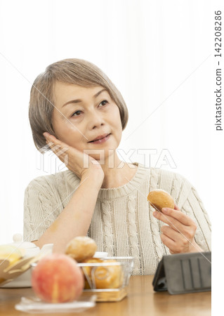 一位老婦人在客廳一邊吃早餐麵包一邊使用智慧型手機。 一位老婦人在客廳一邊吃早餐麵包一邊使用智慧型手機。 142208286