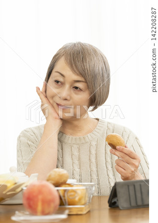 A senior woman eating breakfast bread while using her smartphone in the living room. 142208287