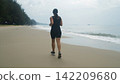 Young woman in black sportswear jogging along a wide sandy beach with lush tropical green trees visible in the background. Nature fitness concept 142209680