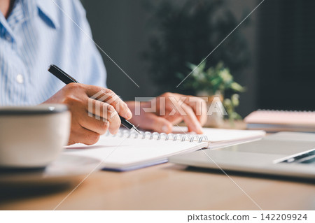 Close-up of a person writing notes in a spiral notebook beside a laptop and coffee cup, representing focused study, remote work, online learning, daily planning, and workplace productivity. Close-up of a person writing notes in a spiral notebook beside a laptop and coffee cup, representing focused study, remote work, online learning, daily planning, and workplace productivity. 142209924