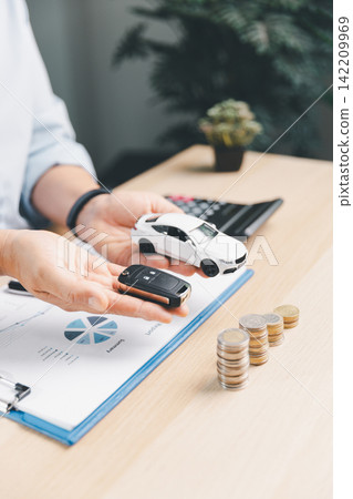 Hands presenting a white toy car and car key over financial report with coin stacks, representing car ownership transfer, auto loan approval, and vehicle insurance. 142209969