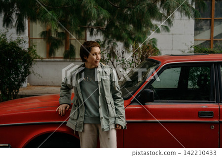 Young looking woman stands next to red car in urban setting during sunny day Young looking woman stands next to red car in urban setting during sunny day 142210433