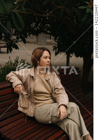 Woman sitting on a bench under a tree in a city park during daytime Woman sitting on a bench under a tree in a city park during daytime 142210447