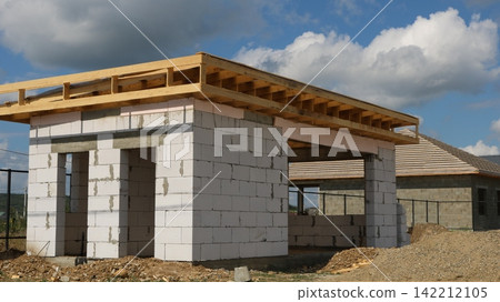 An unfinished one-story building made of white aerated concrete blocks with a wooden frame and a flat roof. Nearby is a neighboring gray house and piles of dirt 142212105