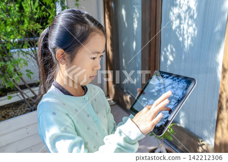 A young elementary school girl operating a tablet on her home balcony. 142212306