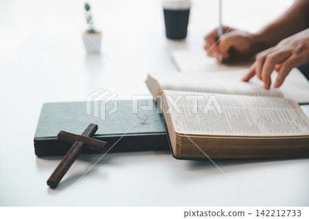 Open Holy Bible with a wooden cross and notebook while a person studies and writes at a bright table, representing Christian learning, devotion, faith, and spiritual reflection. Open Holy Bible with a wooden cross and notebook while a person studies and writes at a bright table, representing Christian learning, devotion, faith, and spiritual reflection. 142212733