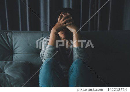 Overwhelmed adult woman sitting alone on a sofa in a dark room with both hands covering her face, expressing stress, anxiety, emotional exhaustion, sadness, and personal crisis. Overwhelmed adult woman sitting alone on a sofa in a dark room with both hands covering her face, expressing stress, anxiety, emotional exhaustion, sadness, and personal crisis. 142212734