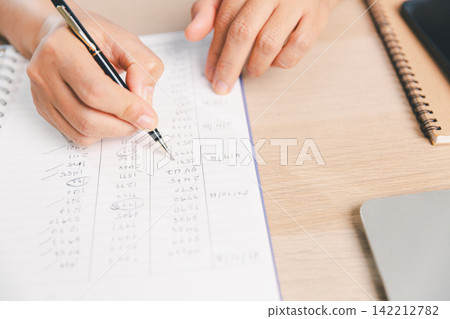 Business professional writing and reviewing financial calculations in a notebook beside laptop, representing accounting, bookkeeping, budgeting, expense tracking, planning, Business professional writing and reviewing financial calculations in a notebook beside laptop, representing accounting, bookkeeping, budgeting, expense tracking, planning, 142212782