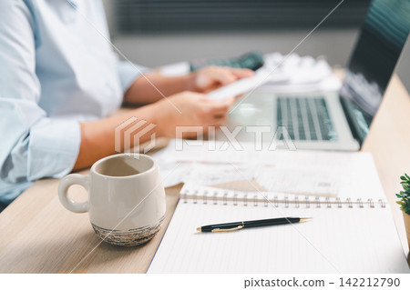 Coffee mug, smartphone, notebook, and laptop on an office desk while a business professional reviews documents, representing productivity, planning, communication, finance, Coffee mug, smartphone, notebook, and laptop on an office desk while a business professional reviews documents, representing productivity, planning, communication, finance, 142212790