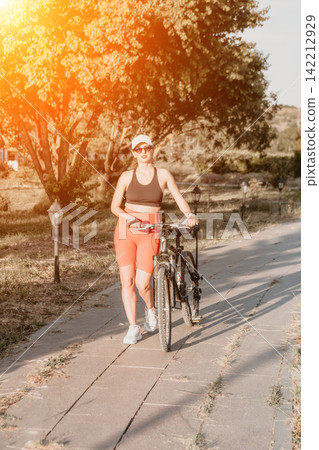 Fitness lifestyle scene featuring athletic woman walking her bicycle along a paved park path during golden hour sunshine Fitness lifestyle scene featuring athletic woman walking her bicycle along a paved park path during golden hour sunshine 142212929
