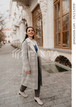 Woman walking city smiles while strolling on a sidewalk wearing a stylish plaid coat and boots in front of architecture Woman walking city smiles while strolling on a sidewalk wearing a stylish plaid coat and boots in front of architecture 142212939