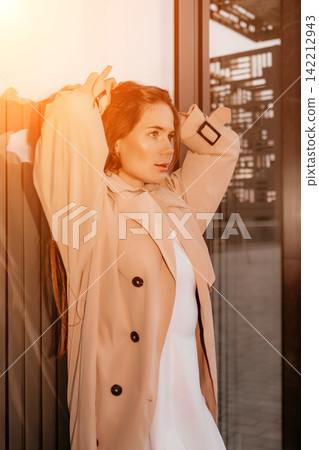 Woman fashion city posing in a beige trench coat against a glass building wall during sunset golden hour 142212943