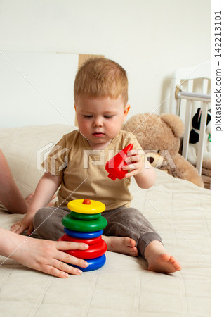 Little boy and mother assembling rainbow pyramid together. Color recognition and size sorting. Creative development through structured toy. Parental support and help Little boy and mother assembling rainbow pyramid together. Color recognition and size sorting. Creative development through structured toy. Parental support and help 142213101