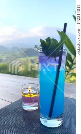 Blue layered cocktail with mint garnish beside glowing candle on wooden terrace table with soft mountain scenery in bright daylight, with cocktail, blue, drink, mint detail Blue layered cocktail with mint garnish beside glowing candle on wooden terrace table with soft mountain scenery in bright daylight, with cocktail, blue, drink, mint detail 142214812