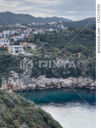 Coastal village nestled in green hills with rocky cliffs and clear blue water background 142215694