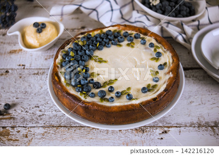 Round cake is topped with cream frosting, blueberries, sitting on wooden table with scattered berries. Round cake is topped with cream frosting, blueberries, sitting on wooden table with scattered berries. 142216120