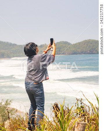 A female tourist crouching on the rocky shore and taking photos with his smartphone at Klayar Beach in Pacitan, East Java, Indonesia. A female tourist crouching on the rocky shore and taking photos with his smartphone at Klayar Beach in Pacitan, East Java, Indonesia. 142217758
