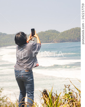 A female tourist crouching on the rocky shore and taking photos with his smartphone at Klayar Beach in Pacitan, East Java, Indonesia. A female tourist crouching on the rocky shore and taking photos with his smartphone at Klayar Beach in Pacitan, East Java, Indonesia. 142217759