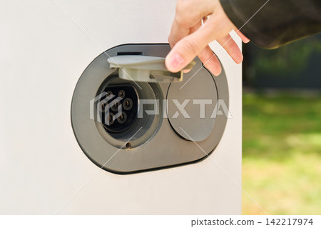 A close-up of an electric vehicle charging port with the protective cover open and a hand preparing the connector for charging. A close-up of an electric vehicle charging port with the protective cover open and a hand preparing the connector for charging. 142217974