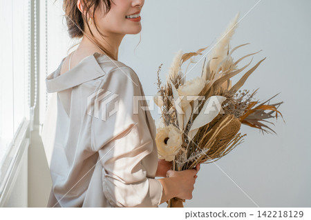 一位衣著時尚的女士手捧乾花，站在客廳窗邊。 142218129