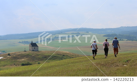 Three hikers walk across a sunlit grassy hill toward a small rural church Three hikers walk across a sunlit grassy hill toward a small rural church 142220213