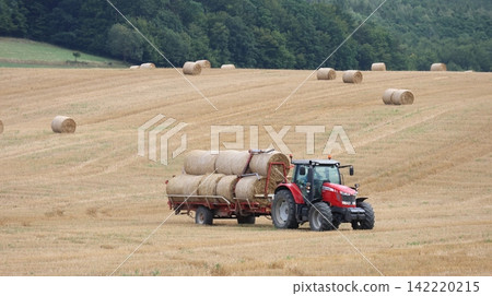 Red tractor towing a trailer loaded with round hay bales across a harvested field Red tractor towing a trailer loaded with round hay bales across a harvested field 142220215
