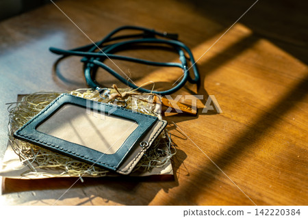 A leather ID card holder is illuminated by diagonal light. Shadows spread across a wooden table. A leather ID card holder is illuminated by diagonal light. Shadows spread across a wooden table. 142220384