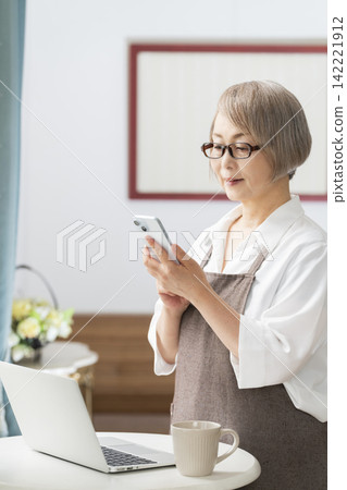 一位老婦人在客廳窗邊使用筆記型電腦。 142221912