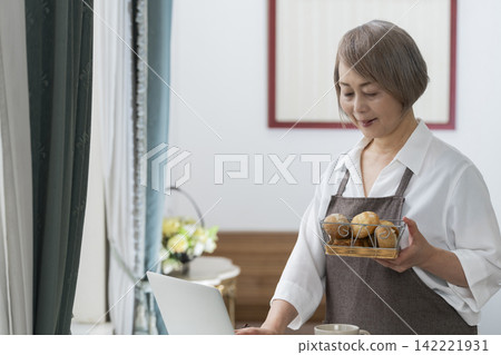 一位老婦人在客廳窗邊使用筆記型電腦。 一位老婦人在客廳窗邊使用筆記型電腦。 142221931