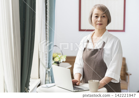 一位老婦人在客廳窗邊使用筆記型電腦。 一位老婦人在客廳窗邊使用筆記型電腦。 142221947