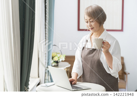 一位老婦人在客廳窗邊使用筆記型電腦。 一位老婦人在客廳窗邊使用筆記型電腦。 142221948