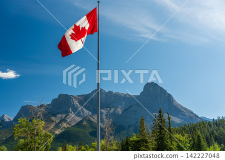 A Canadian flag fluttering in the wind against a backdrop of a summer blue sky and Mount Haring. Canmore, Alberta, Canada. A Canadian flag fluttering in the wind against a backdrop of a summer blue sky and Mount Haring. Canmore, Alberta, Canada. 142227048