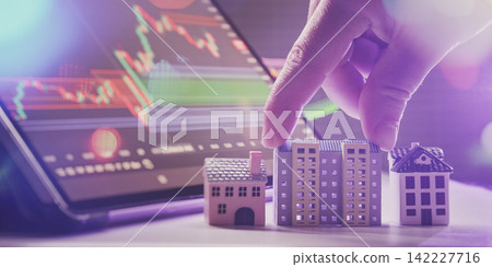 Real estate investment portfolio concept. Hand choosing a miniature apartment building model against a background of stock market candlestick charts. Real estate investment portfolio concept. Hand choosing a miniature apartment building model against a background of stock market candlestick charts. 142227716
