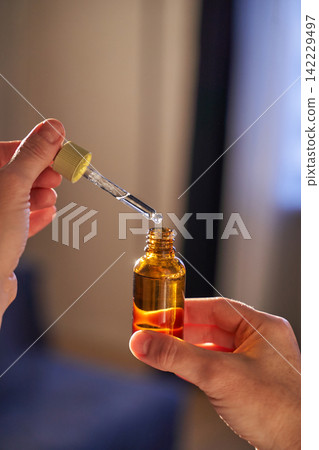 Hands Holding Amber Bottle And Using Dropper To Transfer Oil With Visible Droplet Above Opening Hands Holding Amber Bottle And Using Dropper To Transfer Oil With Visible Droplet Above Opening 142229497