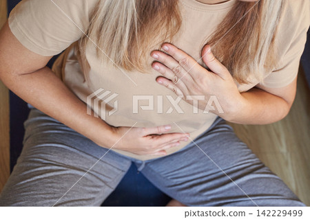 Middle Aged Woman Holding Chest And Abdomen Sitting On Floor Wearing Beige T Shirt And Gray Pants Middle Aged Woman Holding Chest And Abdomen Sitting On Floor Wearing Beige T Shirt And Gray Pants 142229499