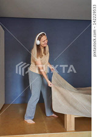 Woman Wearing White Headphones Standing Barefoot Pulling Bedcover Over Mattress In Minimal Bedroom Woman Wearing White Headphones Standing Barefoot Pulling Bedcover Over Mattress In Minimal Bedroom 142229533