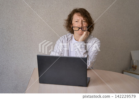 Woman Resting Hand Near Laptop While Looking Tired At Table In Plain Indoor Room With Beige Wall Woman Resting Hand Near Laptop While Looking Tired At Table In Plain Indoor Room With Beige Wall 142229579